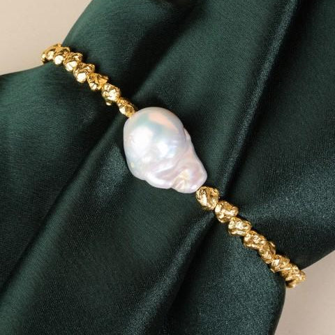 gold plated bracelet around a bunch of dark green shiny fabric. The bracelet has a large gnarly looking pearl in the centre of rugged pebbly shaped beads.
