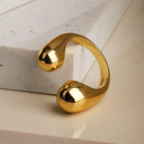 Gold plated ring laying against white marble slab. Two bulbous heads with opening in centre to finger 