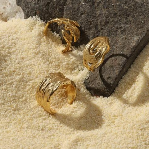 set of three different gold plated textured, organic looking rings sitting in sand near a black rock