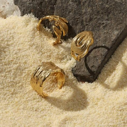 set of three different gold plated textured, organic looking rings sitting in sand near a black rock